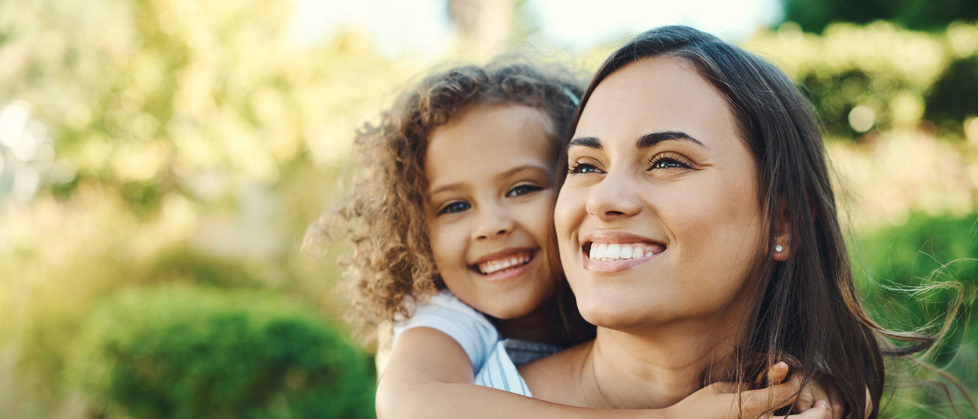 The image shows a woman and a child embracing each other outdoors with trees in the background.